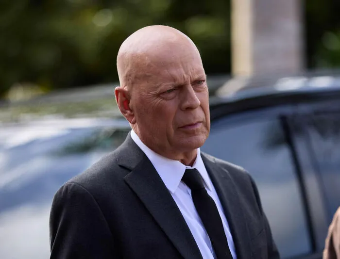 Bald man in a black suit and tie looking serious outdoors near a black vehicle, representing celebrity downfalls.