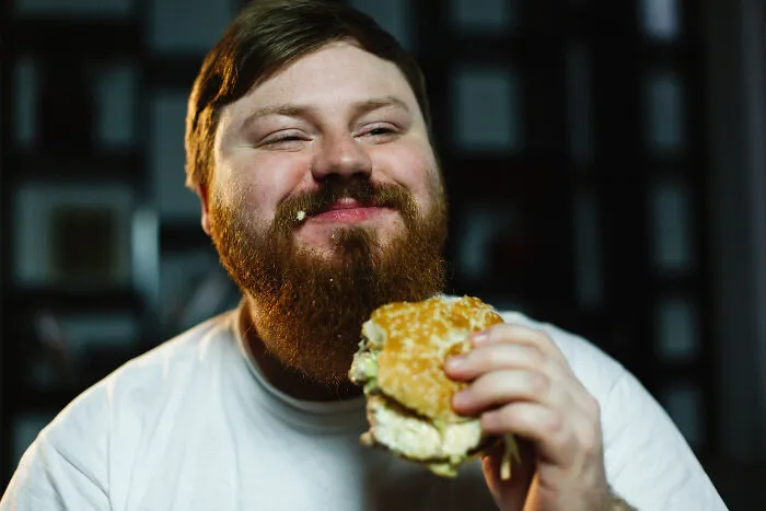 Man with a beard eating a large burger at an all you can eat buffet showing gluttony and indulgence.