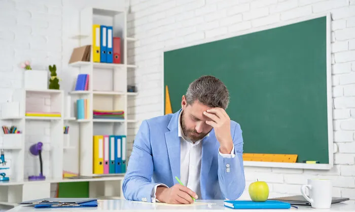 High school secrets: stressed male teacher in classroom writing at desk with chalkboard, apple, books, and mug