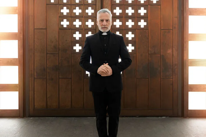 Priest-like older man in clerical collar standing before carved wooden door, high school secrets theme