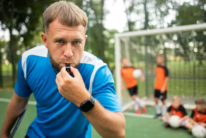 Coach blowing whistle at soccer practice with students by goalpost, high school secrets implied