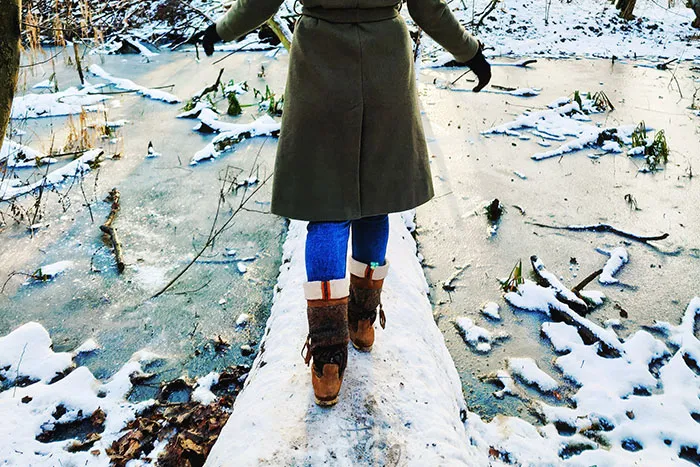 Person wearing brown boots and a coat carefully walking on a snowy log over frozen water, showcasing dangerous things people did.