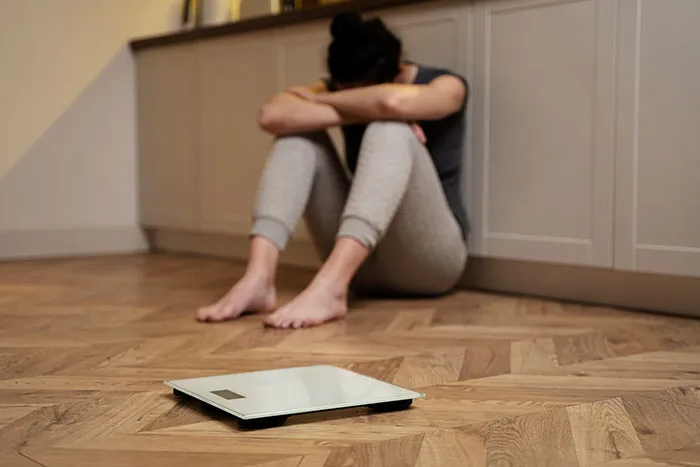Woman sitting on the floor looking distressed near a digital scale, highlighting struggles with women's bodies and body image.
