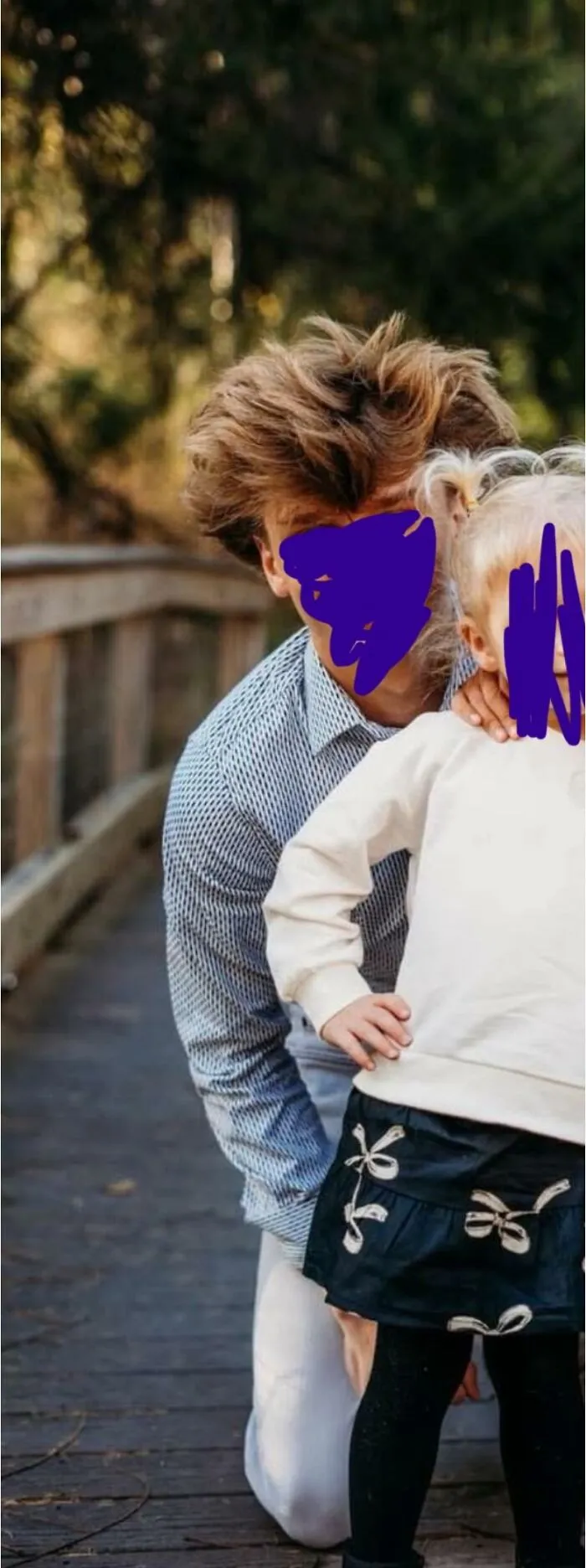 Person with a disastrous hairstyle kneeling outdoors next to a child on a wooden walkway in a natural setting