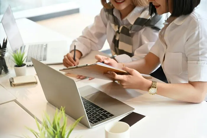 Two coworkers discussing documents and working on laptops in an office, illustrating worst coworker interactions at work.