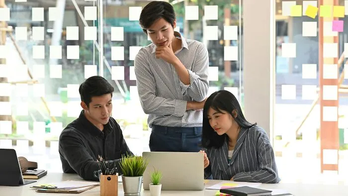 Three coworkers collaborating at a desk with a laptop, surrounded by sticky notes, illustrating worst coworker stories.