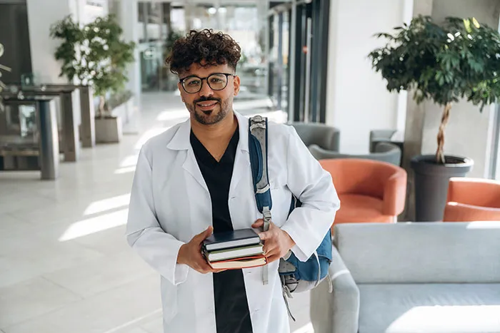 Young gifted man in a white coat holding books, smiling confidently in a modern, bright indoor setting.