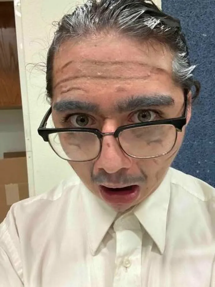 Person wearing stage makeup with exaggerated wrinkles and glasses, creating a magical yet cursed appearance on stage.