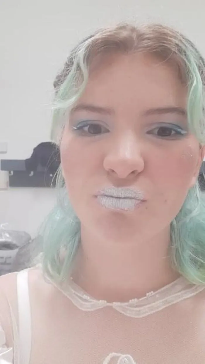 Young woman with light blue stage makeup and glittery silver lips posing for a selfie with a neutral background.
