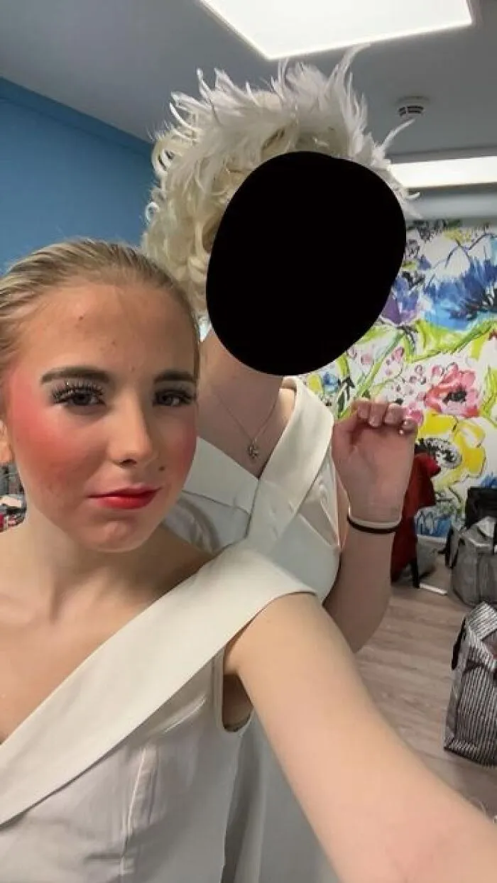 Two performers backstage showcasing stage makeup with exaggerated blush and lashes, dressed in white costumes.