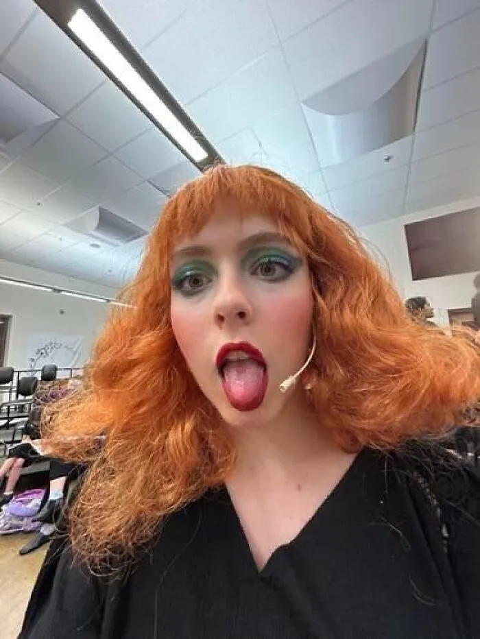 Young woman with bright red hair and exaggerated stage makeup sticking her tongue out in a backstage dressing room.