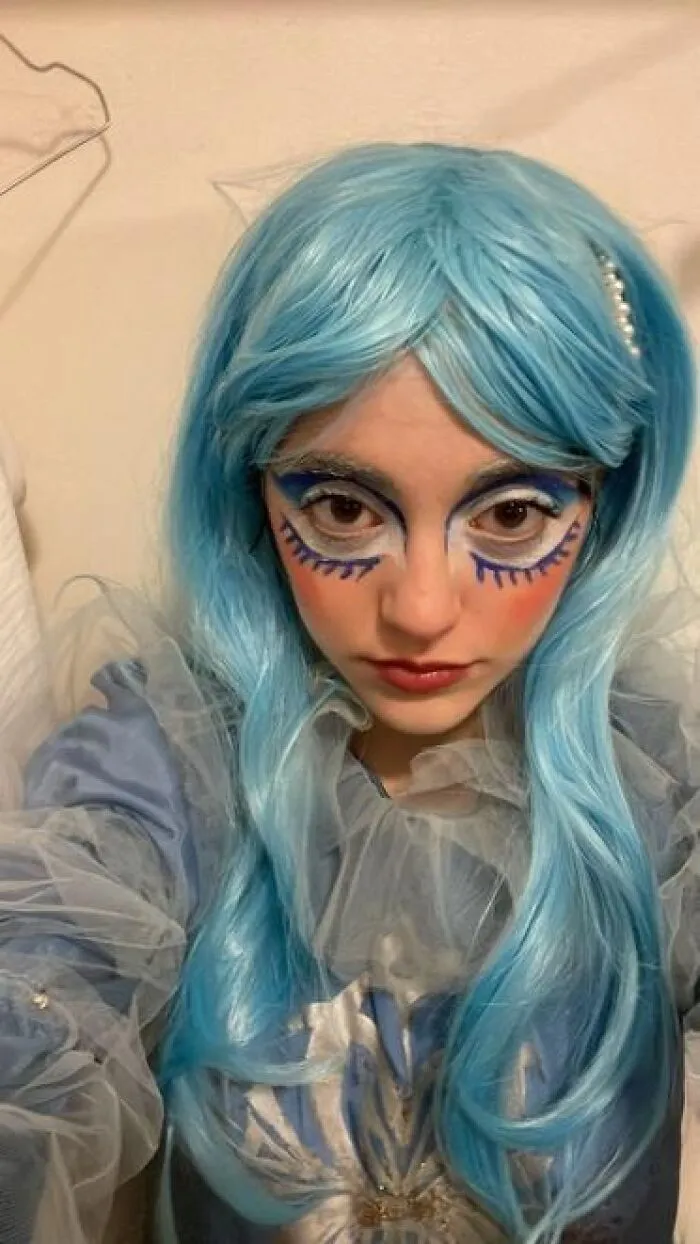 Person with blue wig and dramatic stage makeup featuring bold lashes and white accents in a theatrical costume.