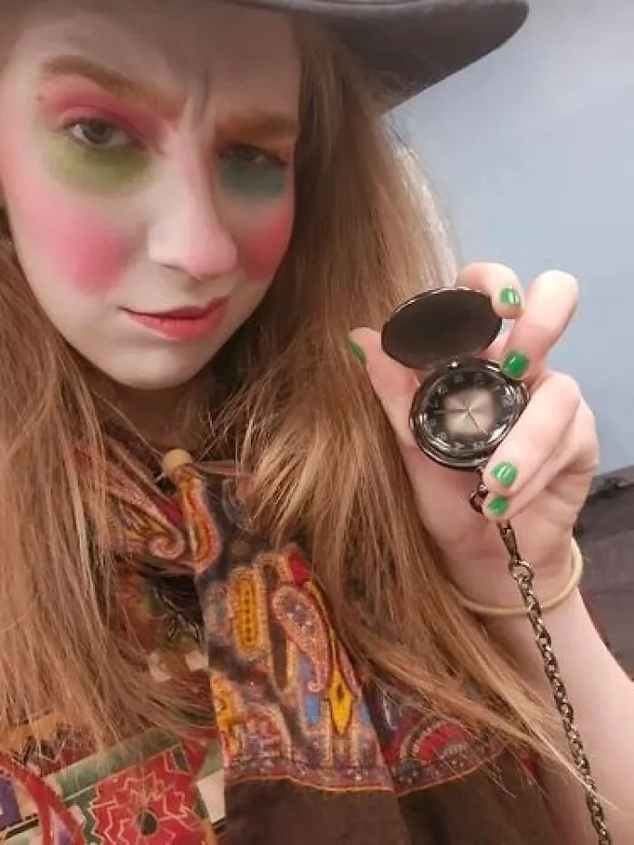Person with stage makeup featuring colorful eyeshadow and blush, holding a vintage pocket watch on a chain.