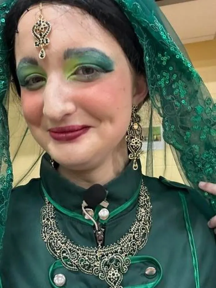 Woman with stage makeup featuring bold green eyeshadow and dark red lipstick, wearing ornate jewelry and a green costume headpiece.