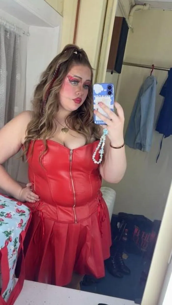 Young woman wearing red dress and dramatic stage makeup taking a mirror selfie in a backstage dressing room.