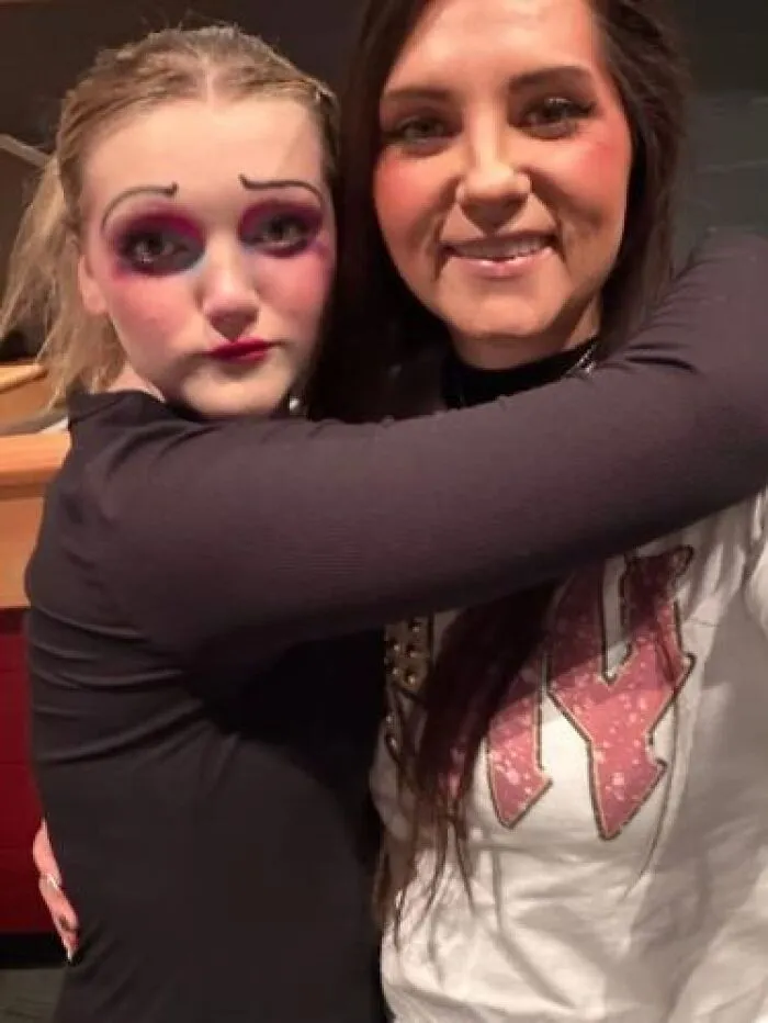 Two women hugging, one with theatrical stage makeup showing an exaggerated, slightly eerie look.