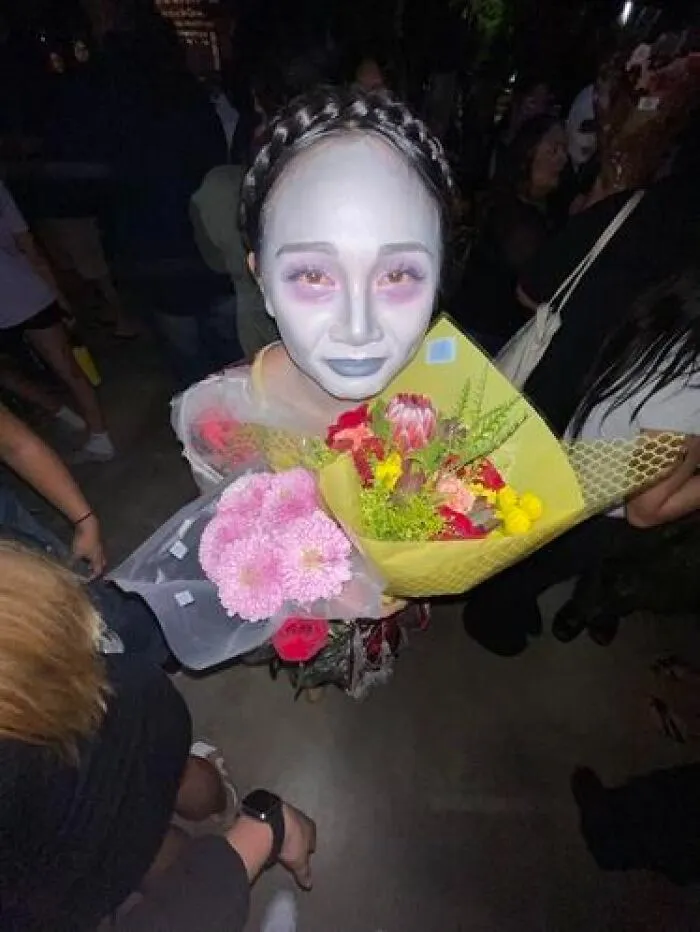 Person with stage makeup in pale face paint and dark lips, holding colorful flower bouquets at a crowded event.