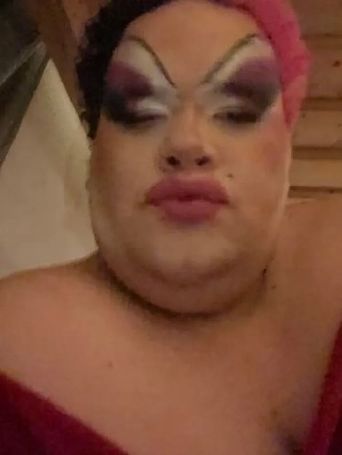Person with bold stage makeup including dark eyeshadow, arched brows, and red lipstick, wearing a pink headband indoors.