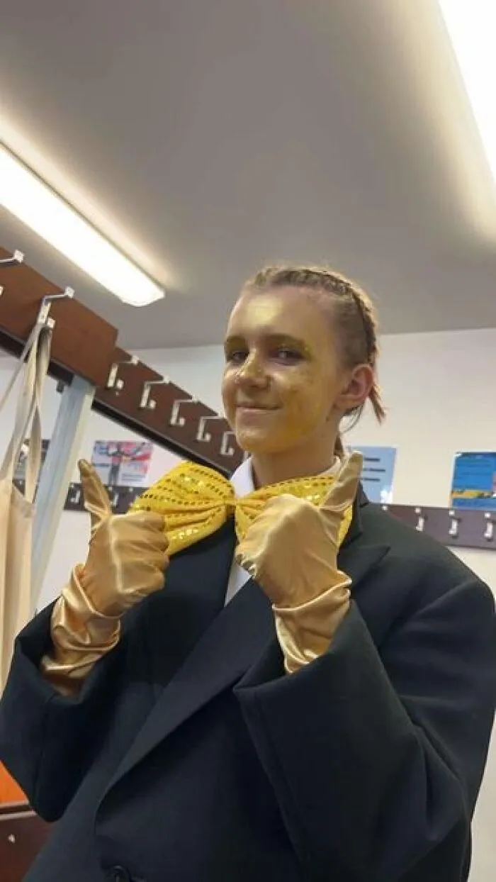 Person with golden stage makeup, wearing gold gloves and a shiny yellow bow tie, posing with thumbs up backstage.