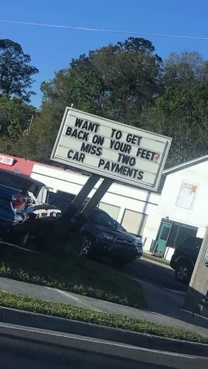 Roadside sign offering help to those who miss two car payments, reflecting themes of dystopia and economic struggle.