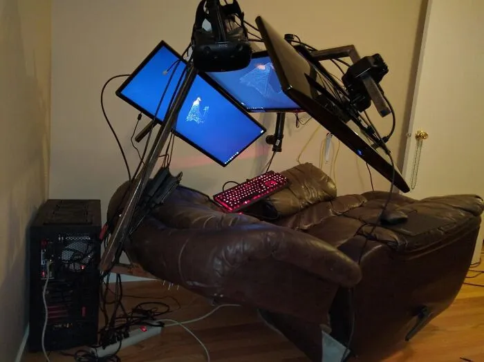 Ergonomic multi-monitor computer setup with VR headset and keyboard attached to recliner, illustrating dystopia technology use.