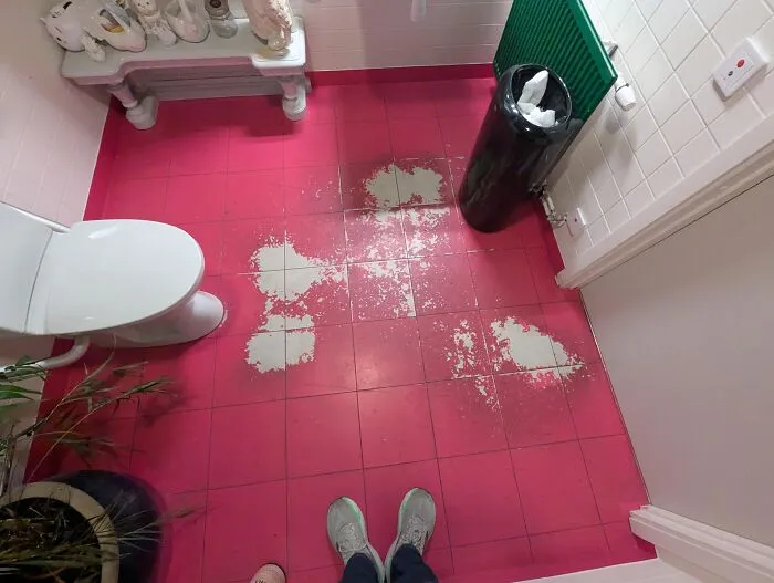 Bathroom floor tiles worn down in unexpected ways with patches of paint missing on red tiles near a white toilet and trash bin.