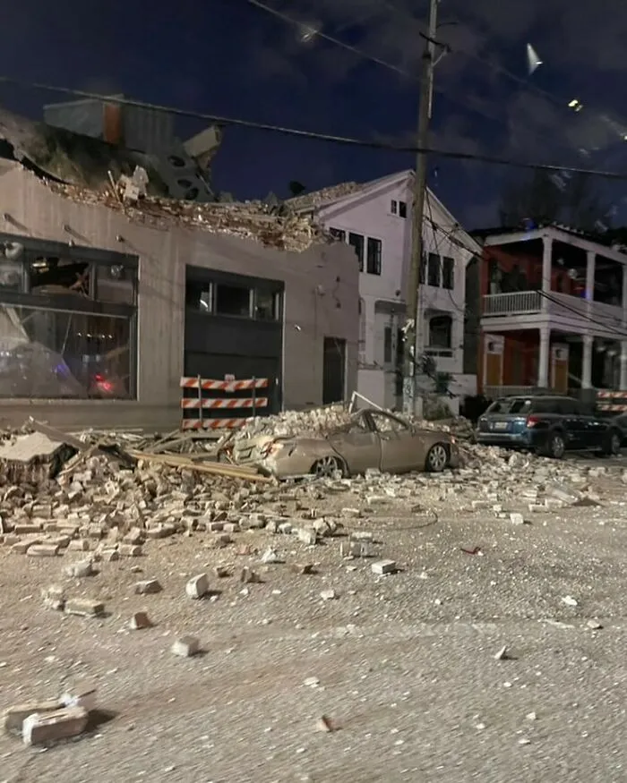 Damaged car covered in bricks and rubble on a street with collapsed building debris in front of houses at night.