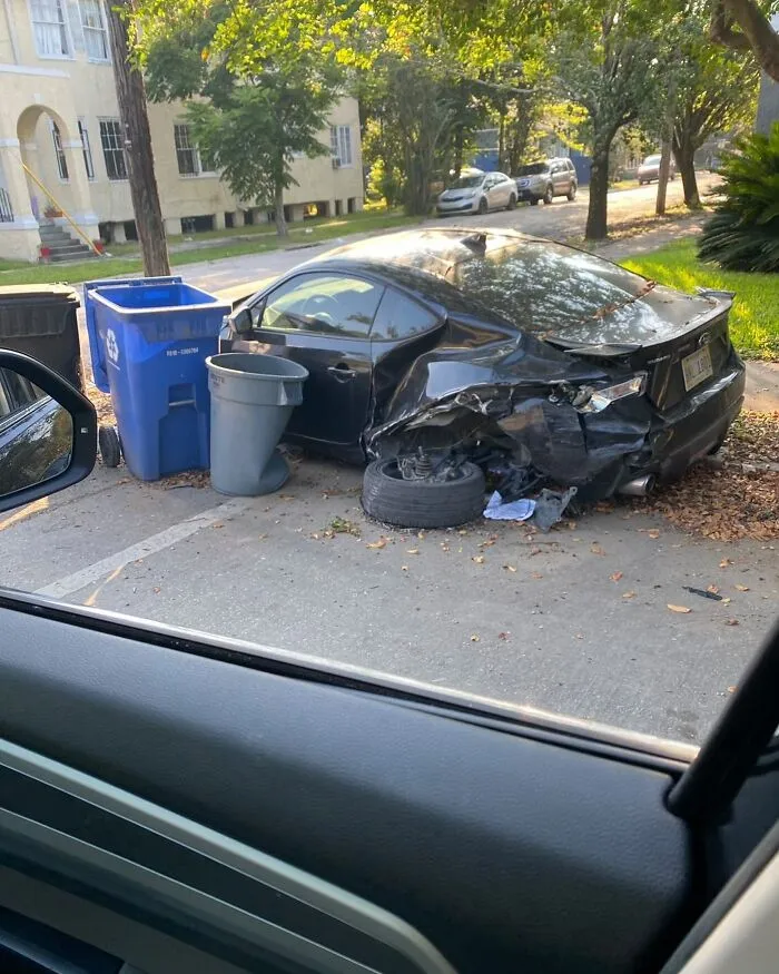 Damaged black car with flat tire parked oddly next to trash bins on a residential street, showcasing weirdest things found in streets.