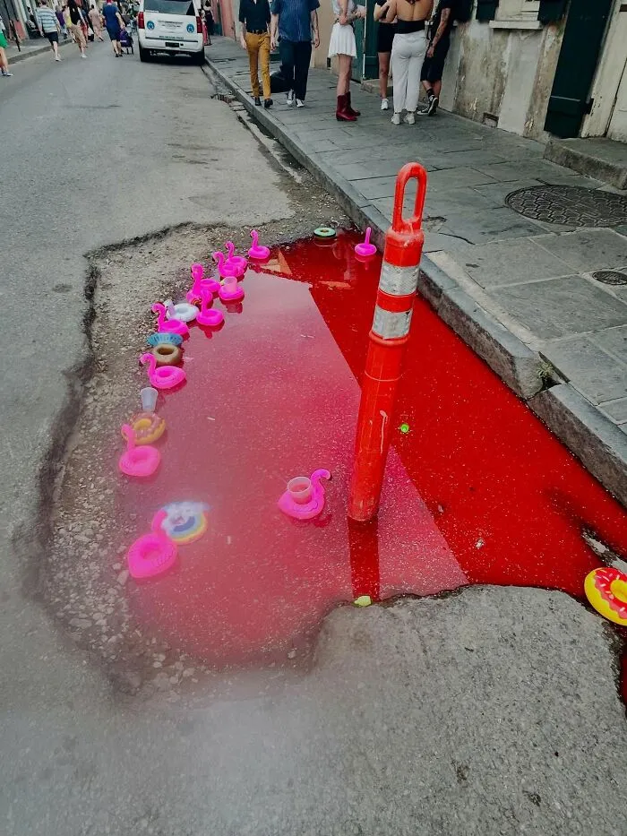 Pothole filled with red water and decorated with pink flamingo floaties and a red traffic cone on the street.
