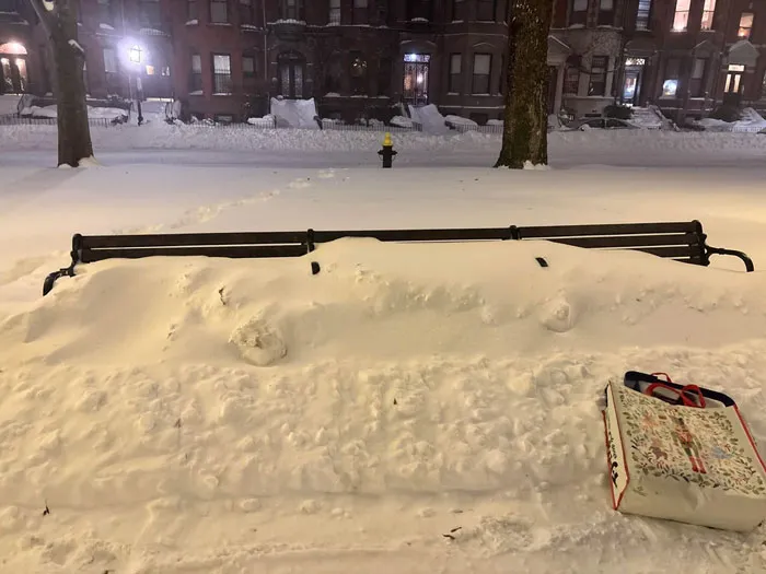 Park bench covered in heavy snow during U.S. snowstorm with buildings and trees in the background at night