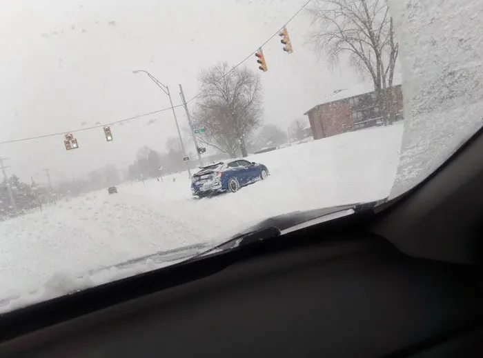 View from inside a car of a snowstorm hitting the U.S., showing heavy snow covering roads and a blue car stuck near an intersection.