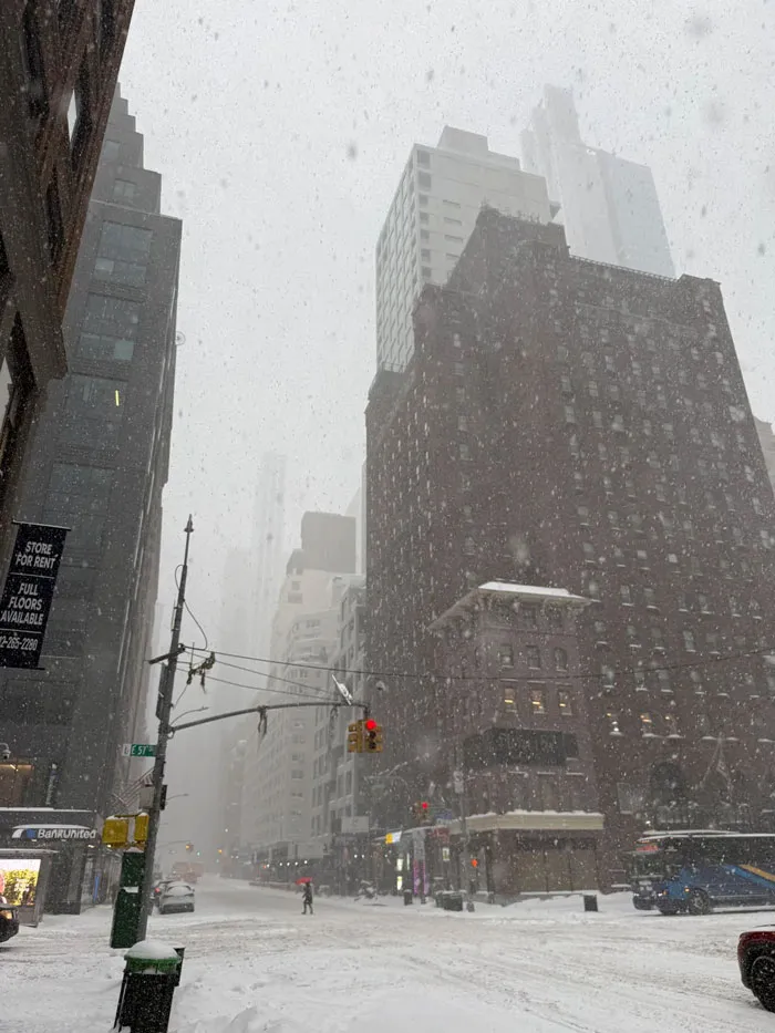 Snowstorm hitting a U.S. city street with heavy snowfall, reduced visibility, and people braving the winter weather.