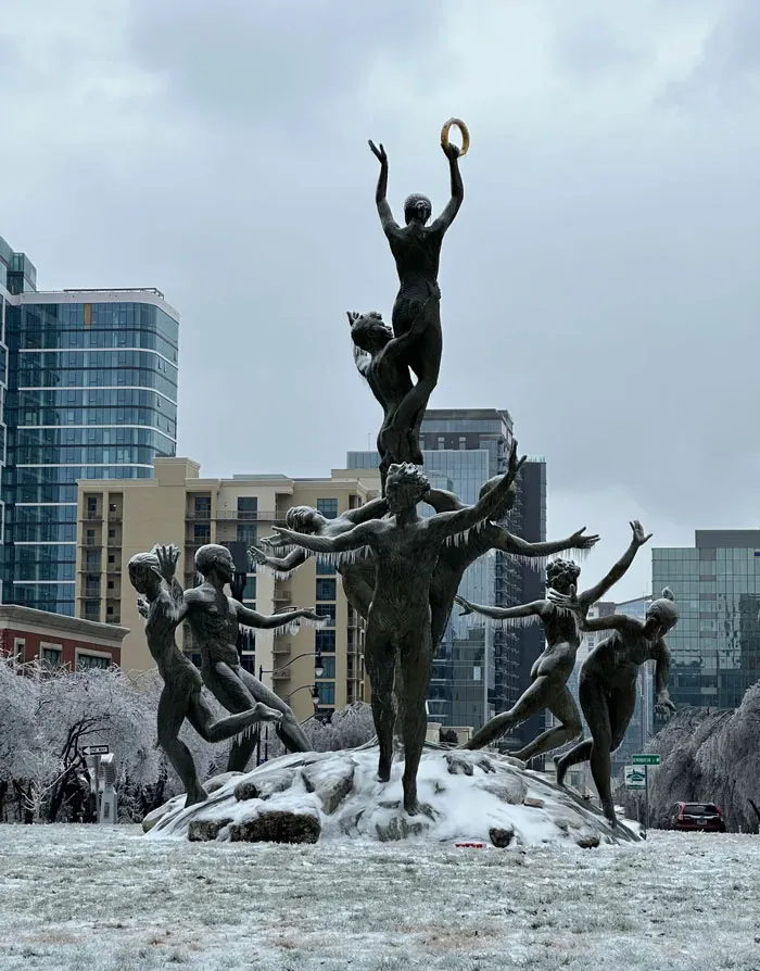 Snowstorm hitting the U.S. with a snowy statue of dancing figures in an urban park on a winter day.