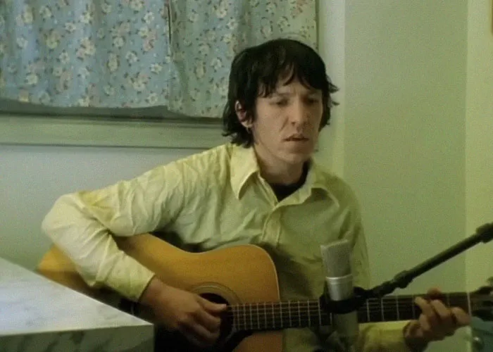 Young man playing acoustic guitar while singing into a microphone, related to celebrity passings topic.