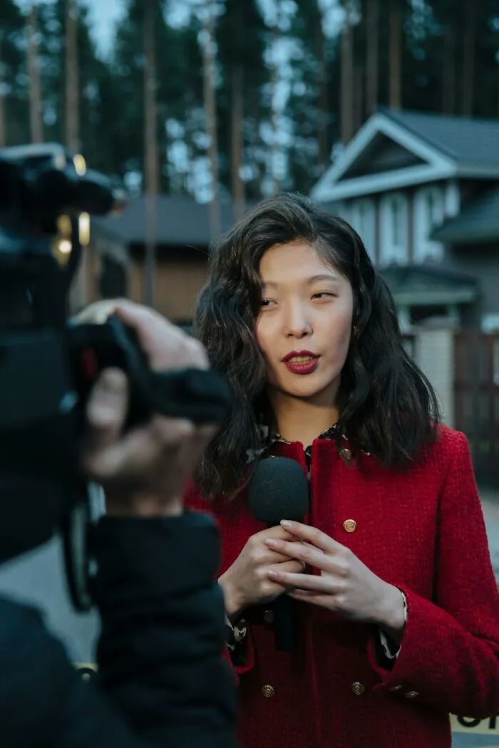 Female reporter holding microphone outside, sharing local inside jokes during a recorded interview in a residential area.