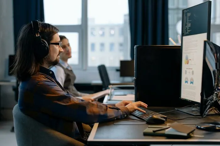 Man wearing headphones working on a computer in an office with coworkers, illustrating worst coworkers ever stories.