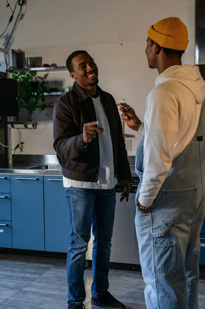 Two men laughing and talking in a kitchen, one wearing a yellow beanie and overalls, rained frogs
