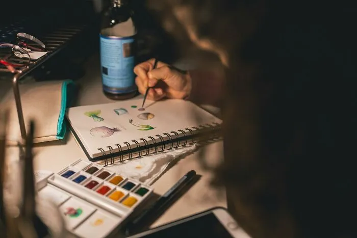 Person painting with watercolors in a sketchbook, creating hopeful and uplifting artwork symbolizing progress in America.