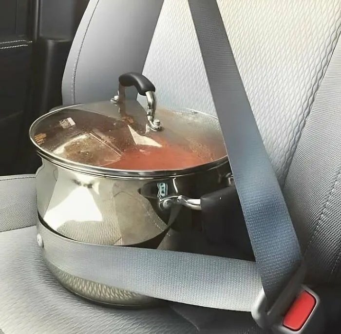 Large cooking pot with lid filled with food secured by a seatbelt on a car seat, capturing Eastern European experience humor.