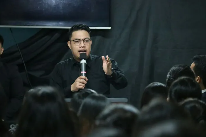 Man speaking into a microphone to an audience, sharing experiences of former cult members in a dark room setting.