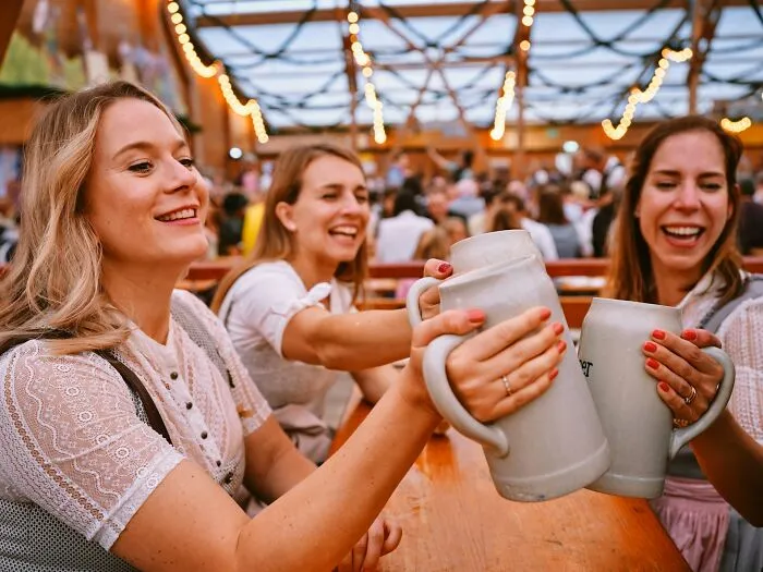 Three women clinking large beer steins and enjoying time at a lively tourist attraction with string lights overhead.