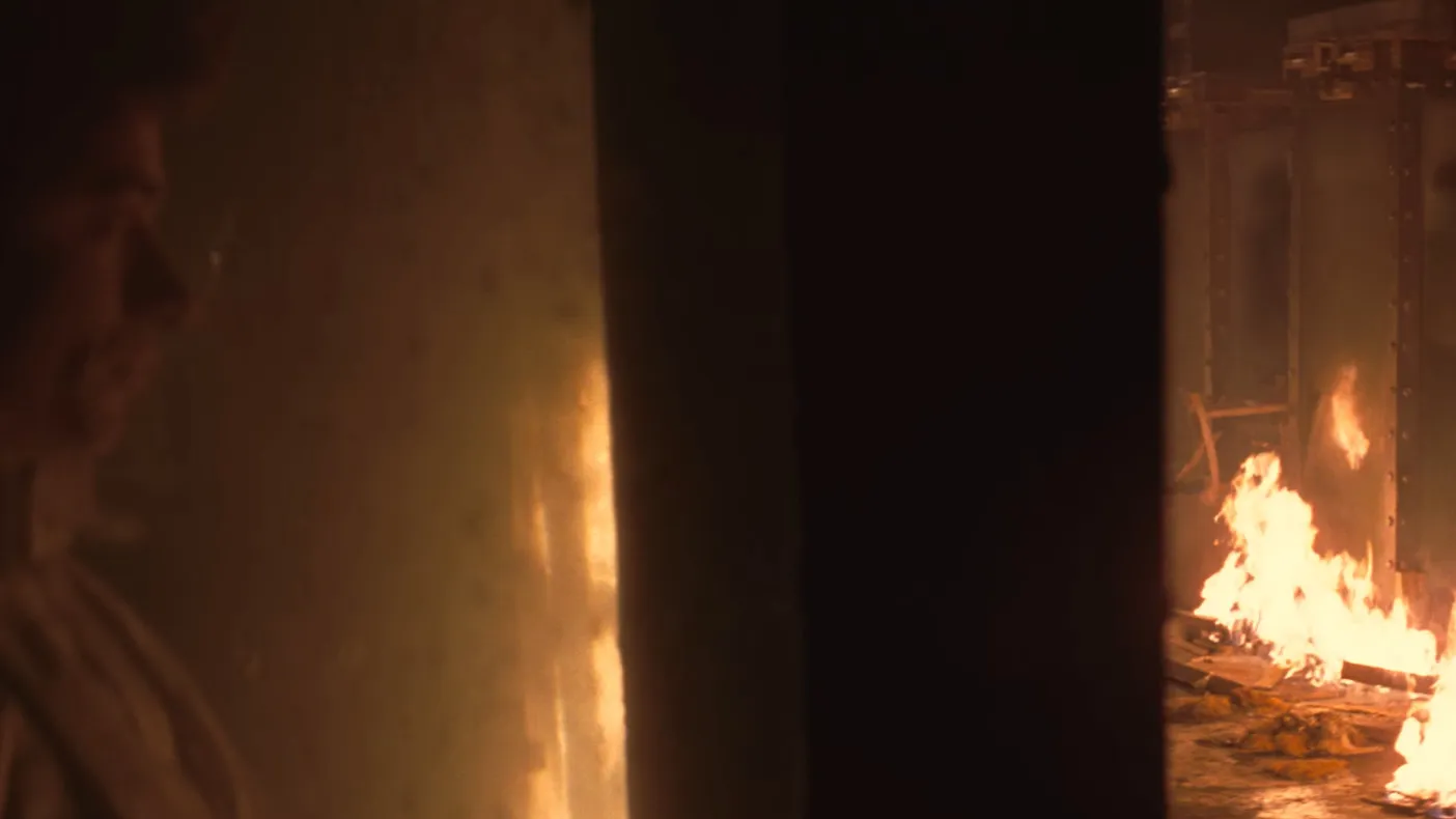 Man partially visible behind wall with fire blazing in a dimly lit room, depicting a movie final scene moment.