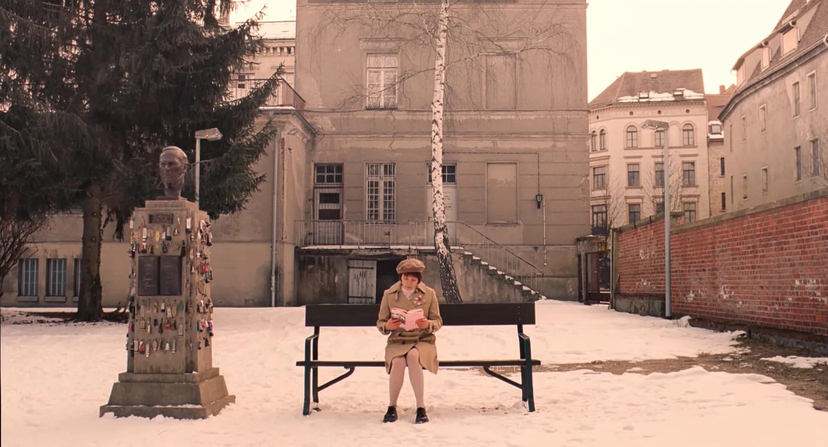 Woman reading a book on a bench in snow near a monument and old buildings in a movie final scenes setting.
