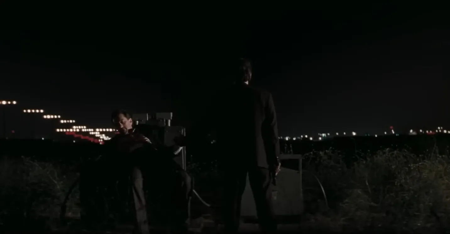 Two men in a dark outdoor setting during a tense movie final scene at night with city lights in the background.