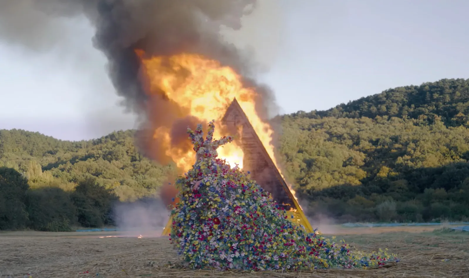 A colorful flower-covered figure in front of a burning pyramid, depicting a dramatic movie final scene.
