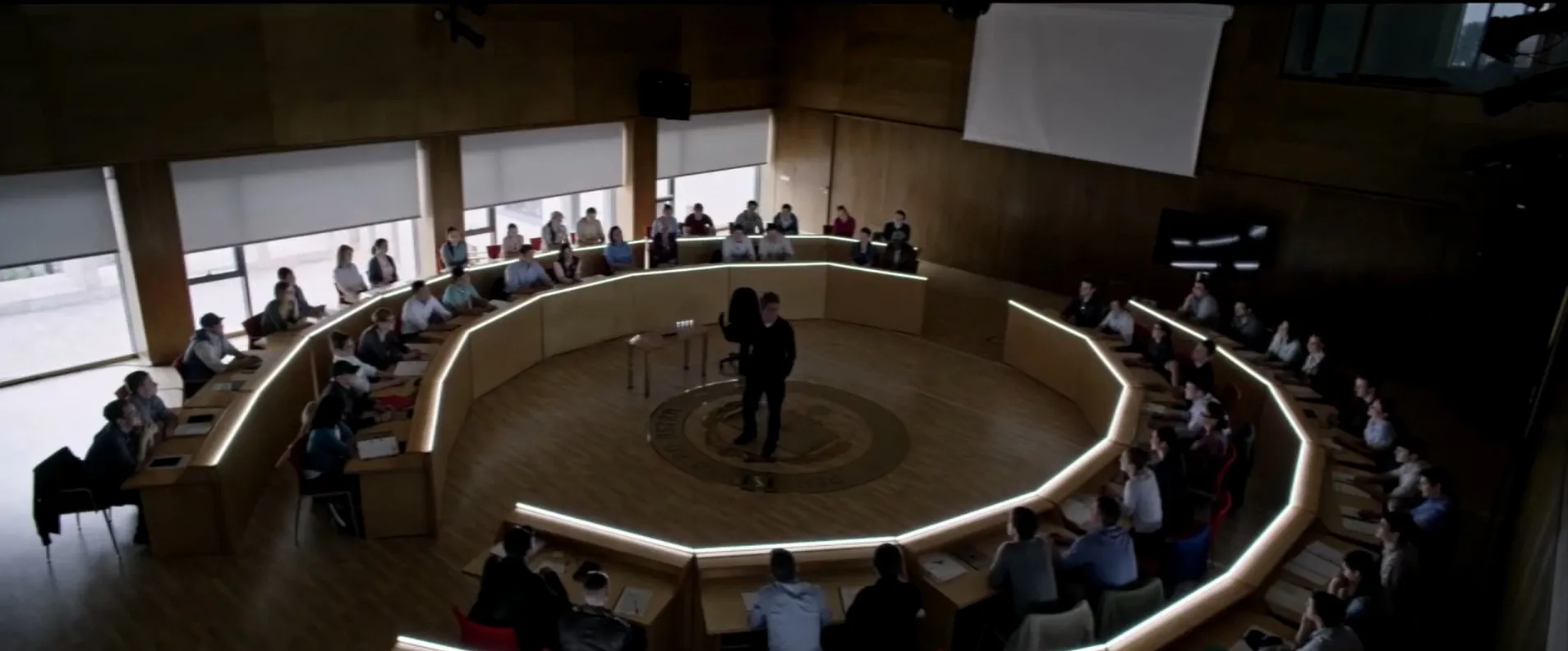 A man stands in the center of a large circular conference room surrounded by seated people in a movie final scenes setting.