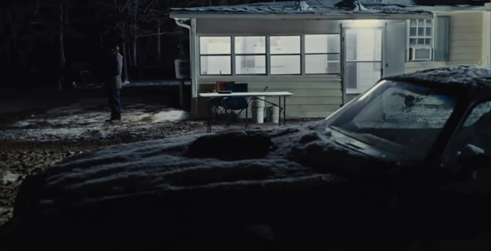 Dark night scene showing a snowy vintage car parked outside a lit cabin with a person standing nearby, movie final scenes.
