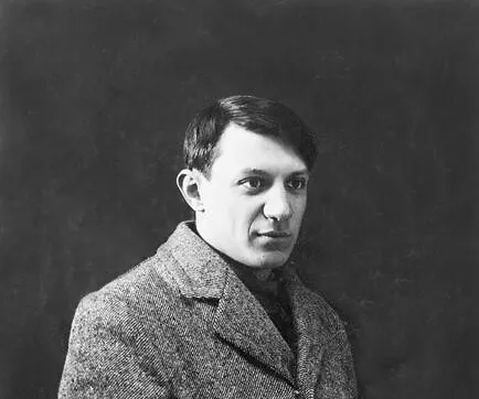 Black and white portrait of Spanish painter Pablo Picasso wearing a coat against a dark background.
