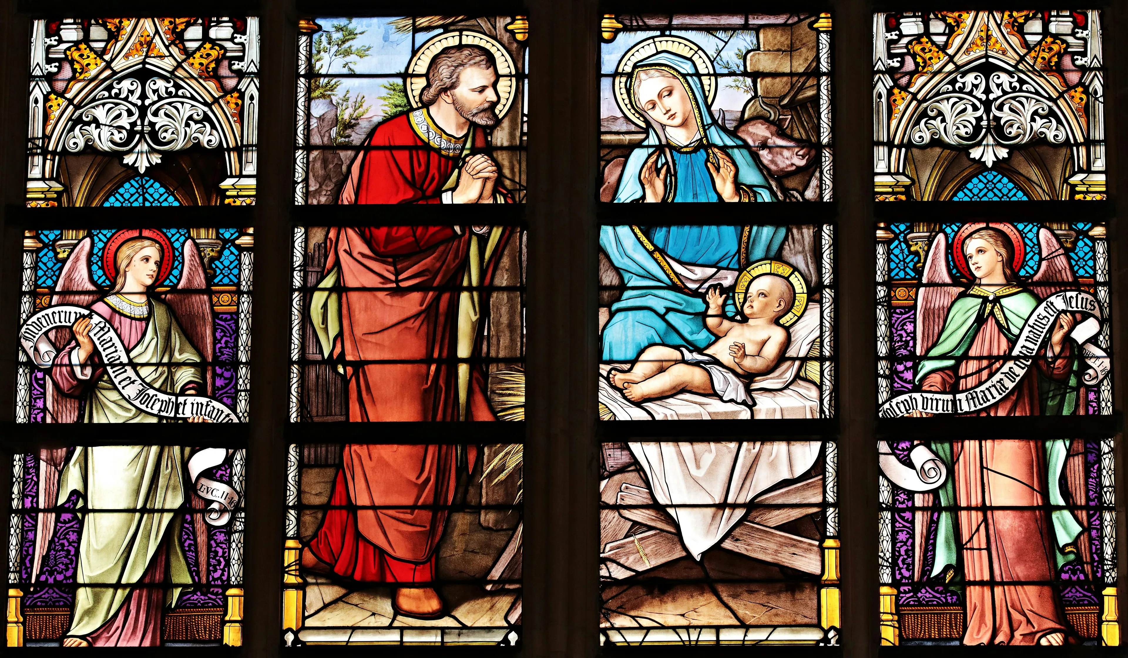 Medieval cathedral stained glass artwork depicting a biblical scene with Mary, baby Jesus, and angels glowing in sunlight.