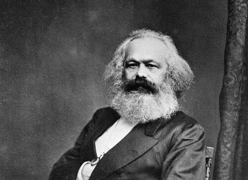 Portrait of Karl Marx with white hair and beard, seated and wearing a dark suit in a vintage black and white photo